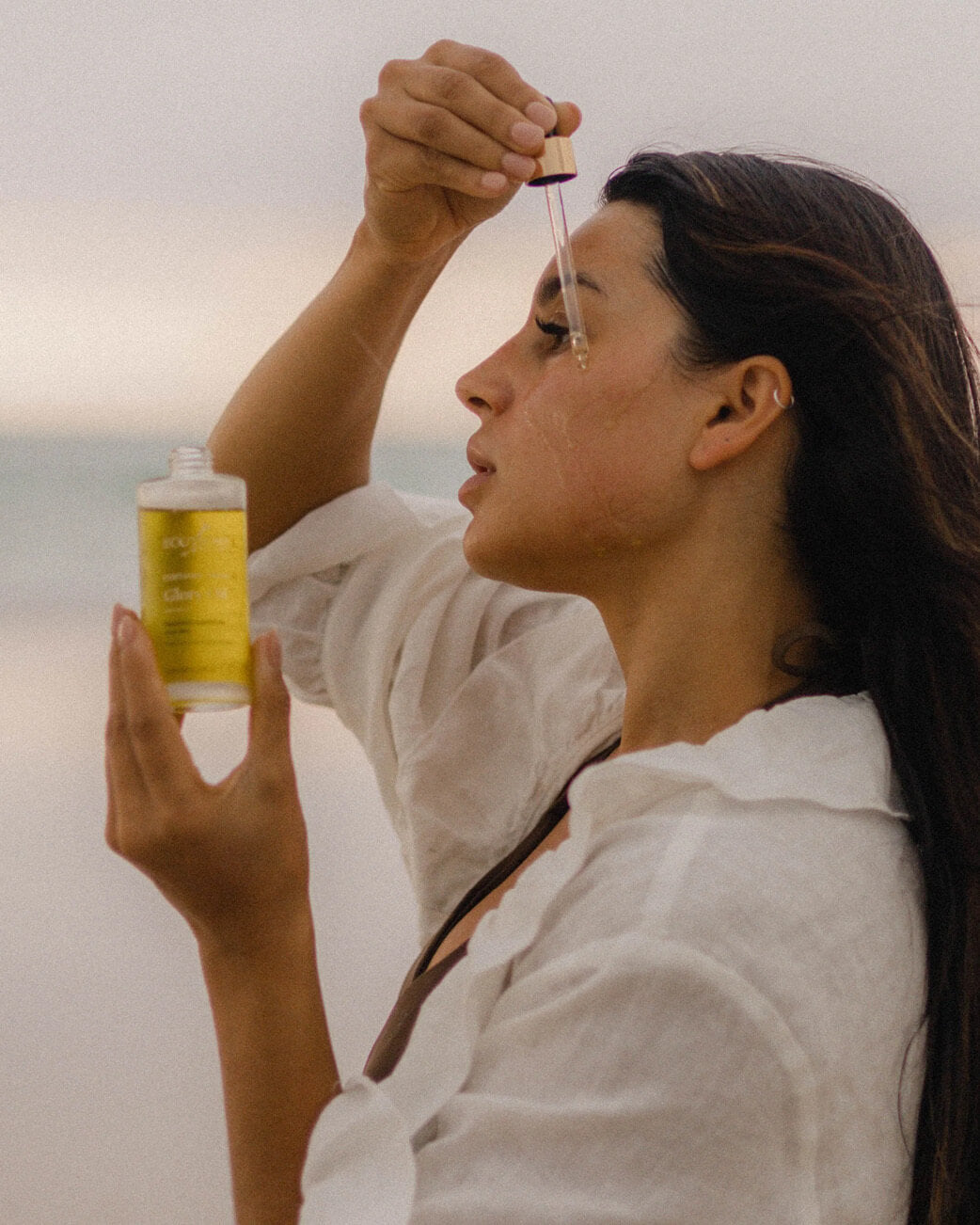 Woman applying a dropper of oil to her face with a bottle of oil in the foreground.