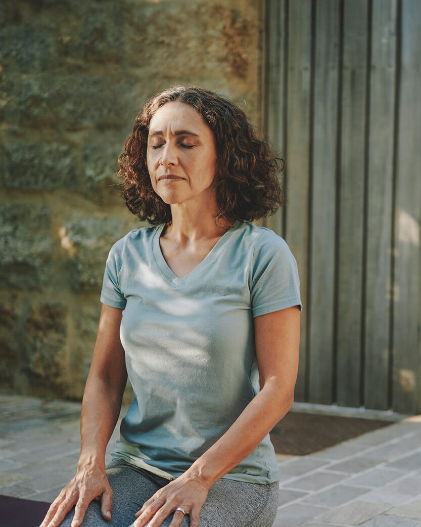Woman sitting on a yoga mat outdoors with a stone wall in the background.