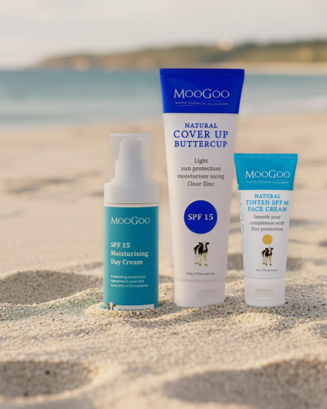 Three Moogoo sunscreen products on a sandy beach with water in the background.