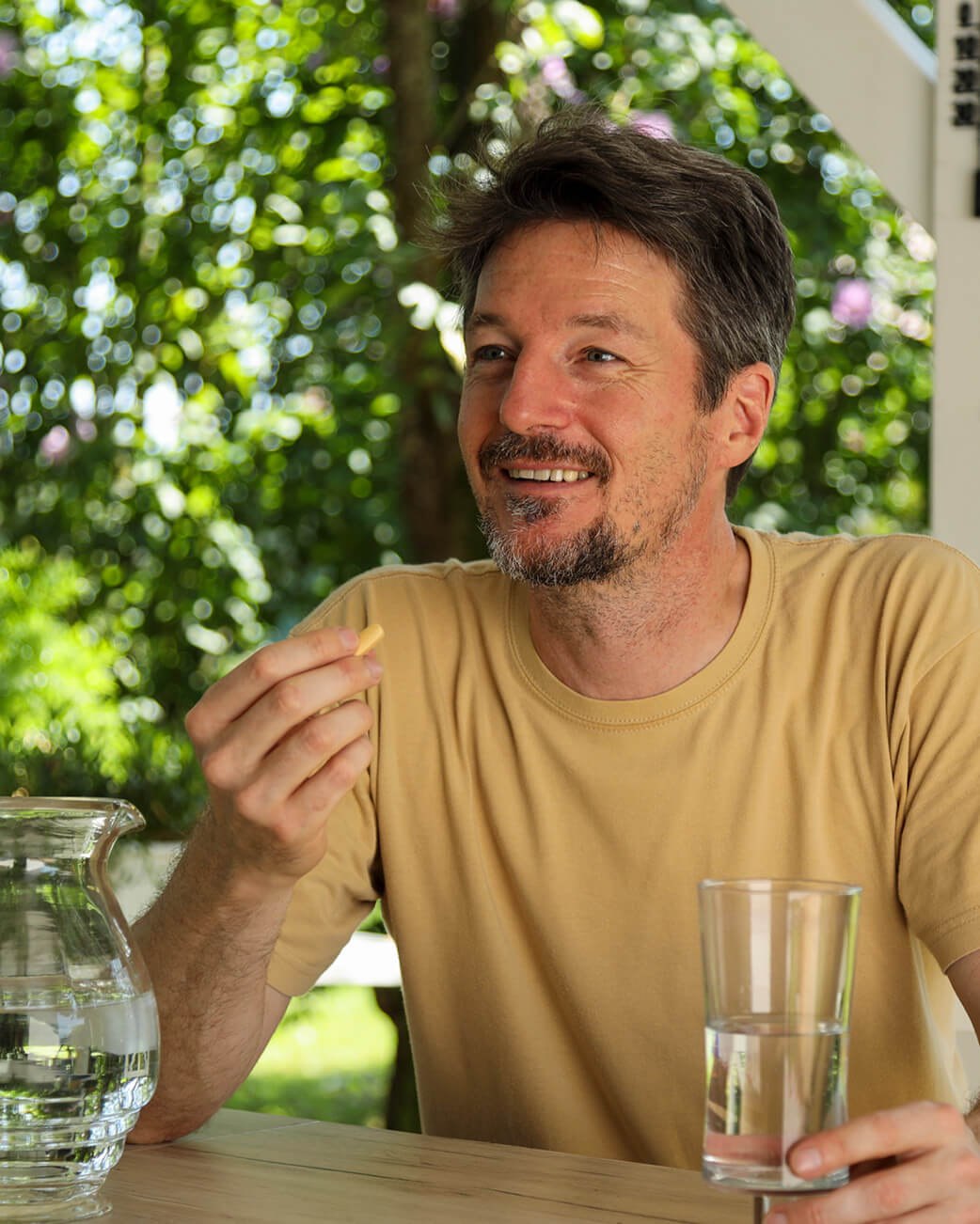 Man sitting outdoors, holding a glass of water and a supplement capsule, with greenery in the background.