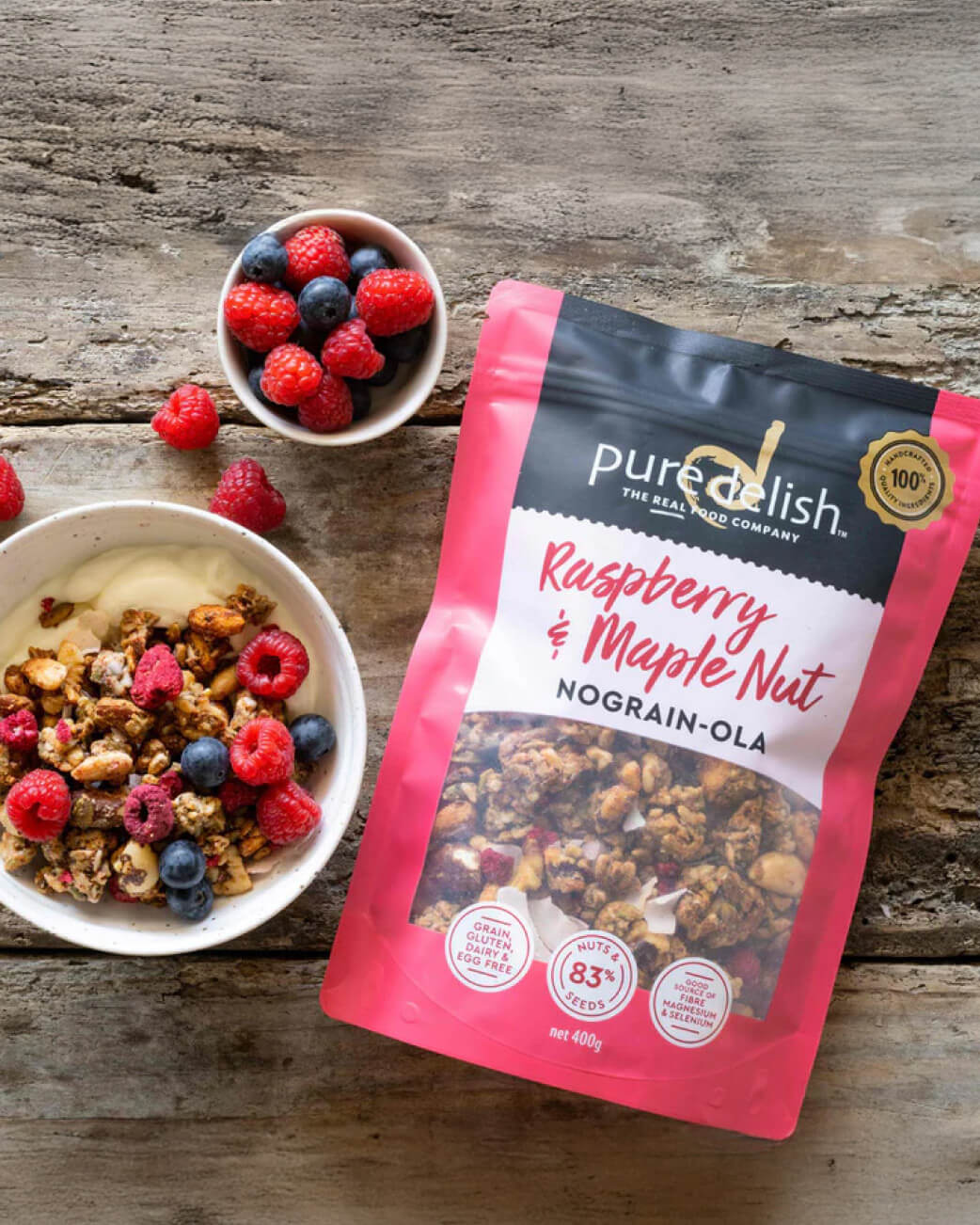 Bag of PureDelish Raspberry & Maple Nut Nograin-Ola on a wooden surface with a bowl of granola and berries.