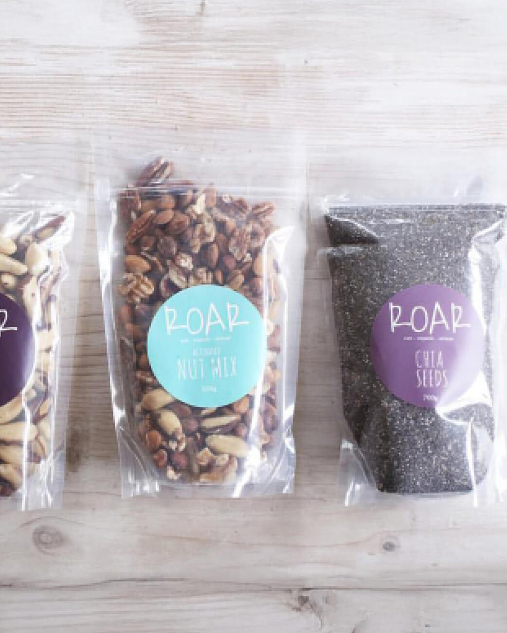 Three packages of roasted nuts and seeds with 'ROAR' branding on a wooden surface.