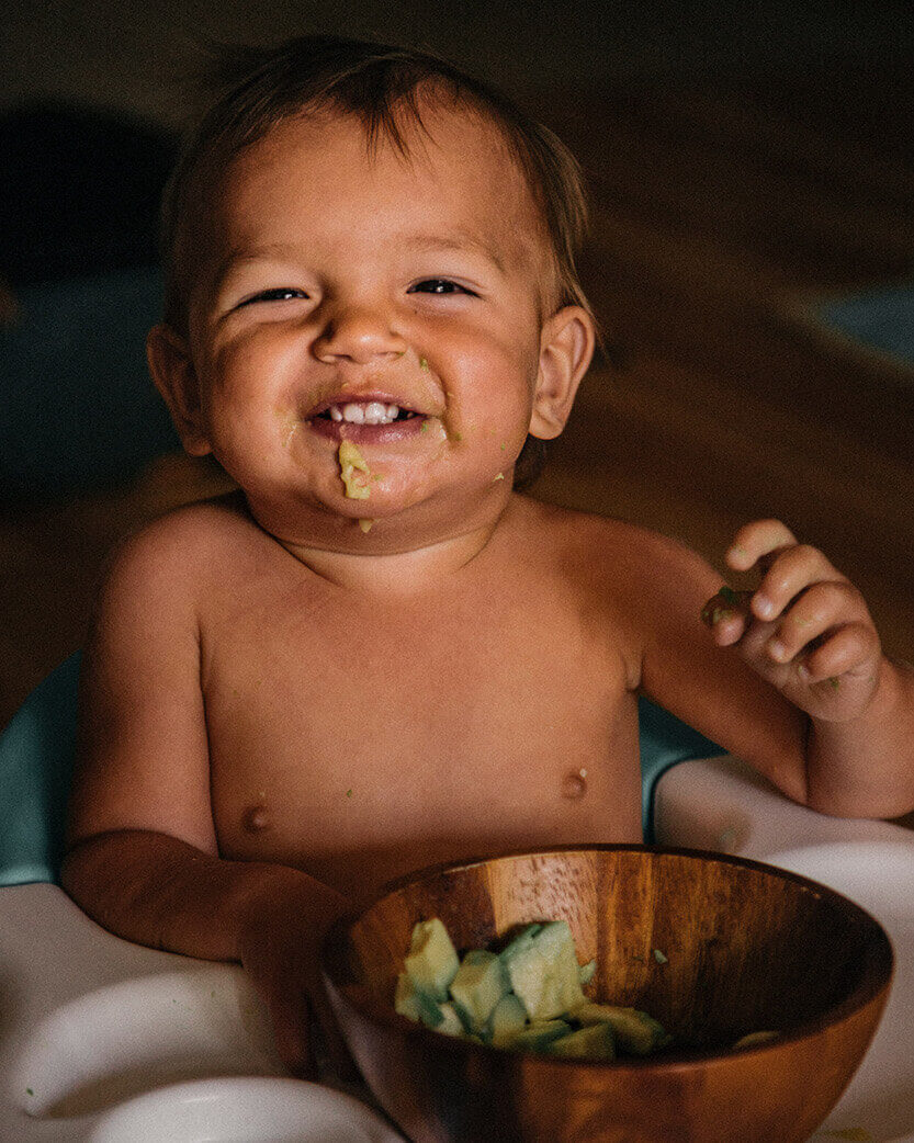Baby eating from a wooden bowl with food on a dark background.