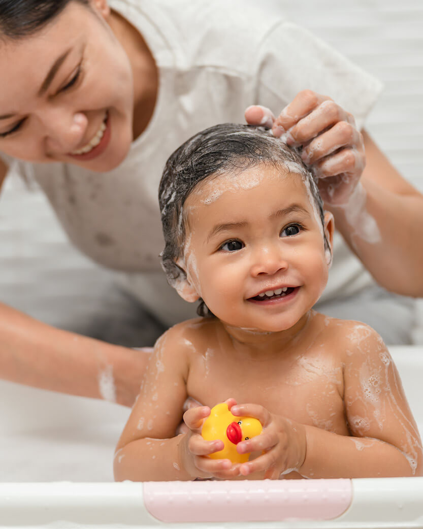 Woman washing a child's hair with a yellow rubber duck in the bathtub.