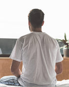 Man sitting on a bed wearing a white t-shirt, looking out of a window.