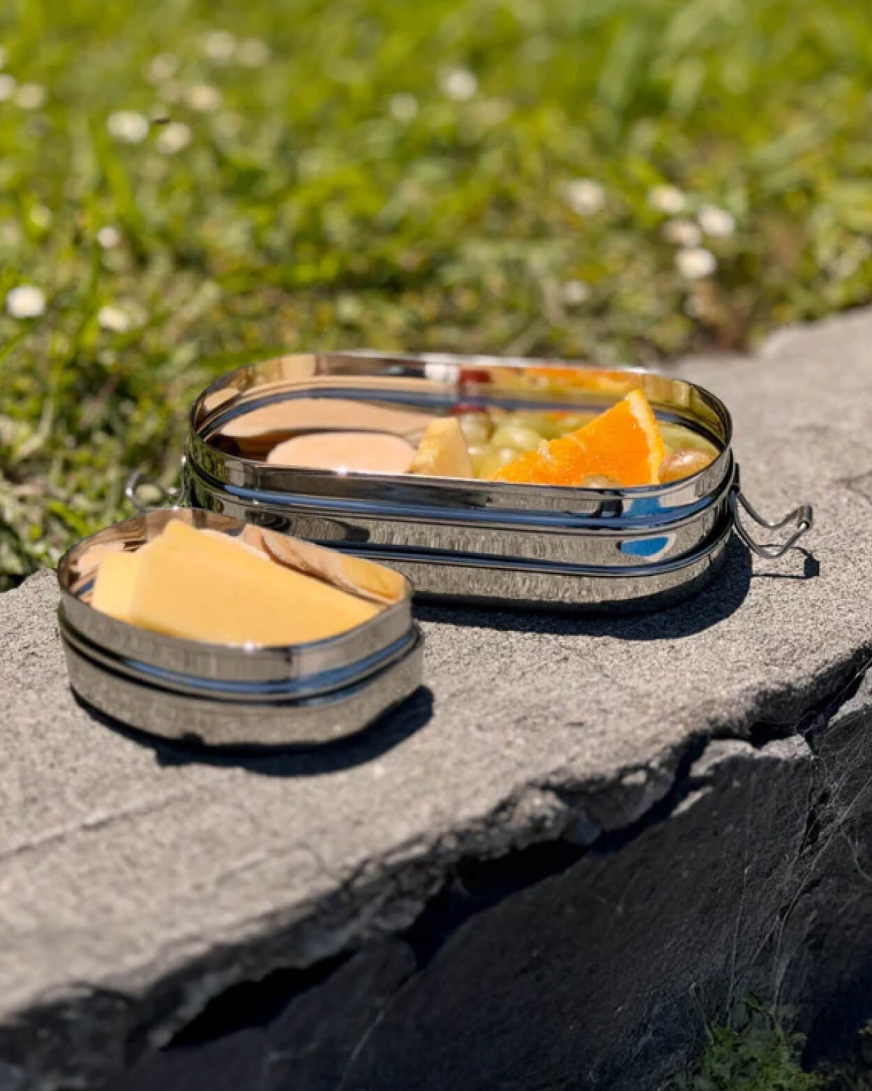 Two metal lunch containers with food on a stone surface outdoors.