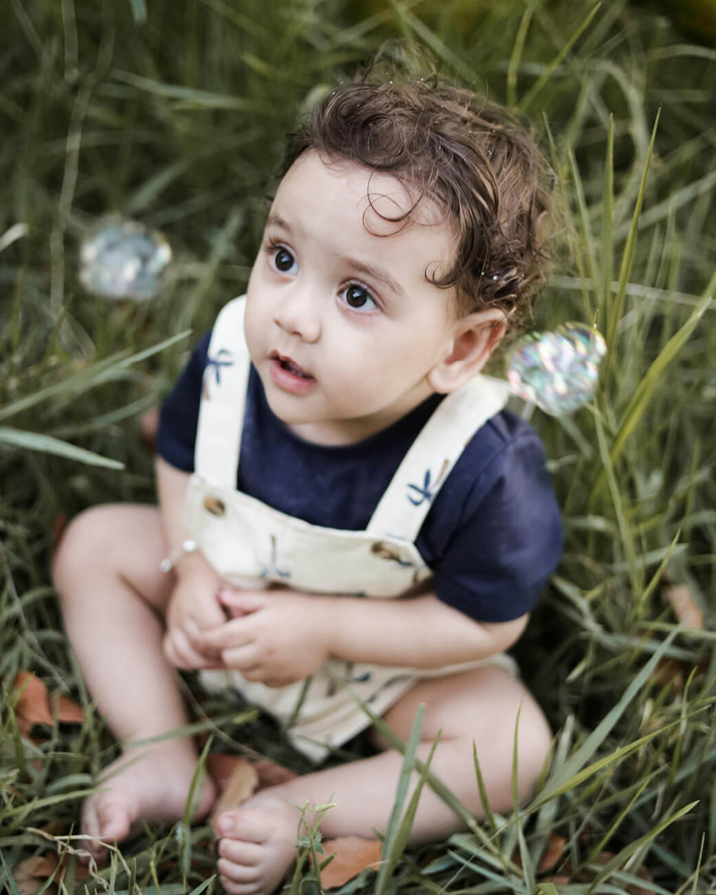 Child sitting in grass wearing a navy shirt and beige overalls.