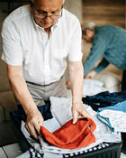 Man folding clothes in a suitcase with another person in the background.
