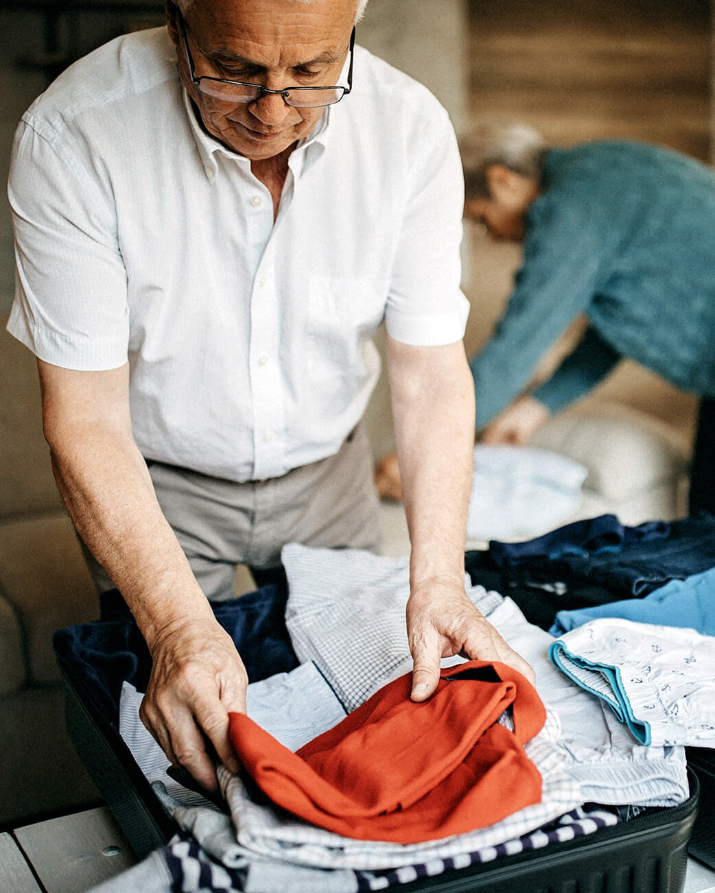 Man folding clothes in a suitcase with another person in the background.