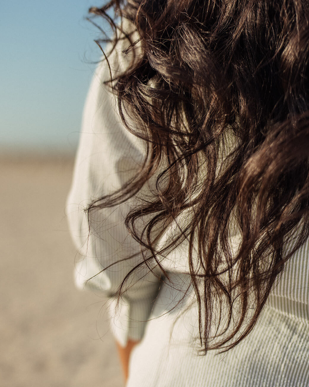 Close-up of a person with wavy hair blowing in the wind, wearing a light-coloured top.