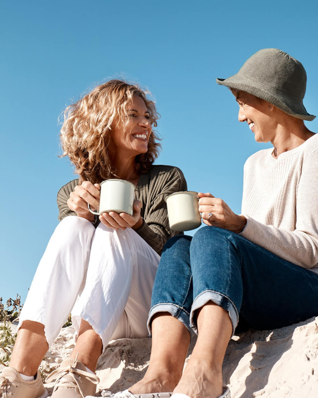 Two people sitting on a beach, holding mugs and smiling at each other.