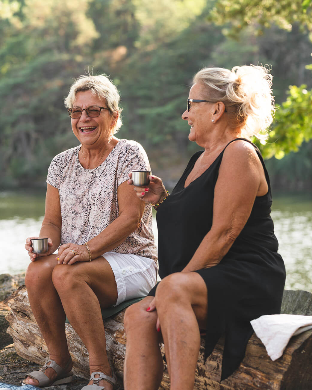 Two women sitting on a log drinking out of cups and laughing in a natural setting.