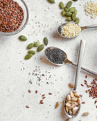 Assorted seeds and grains on a light surface with spoons.