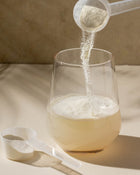 Powder being poured from a scoop into a glass of liquid on a neutral background