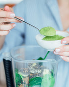 Person adding green powder to a blender with a spoon