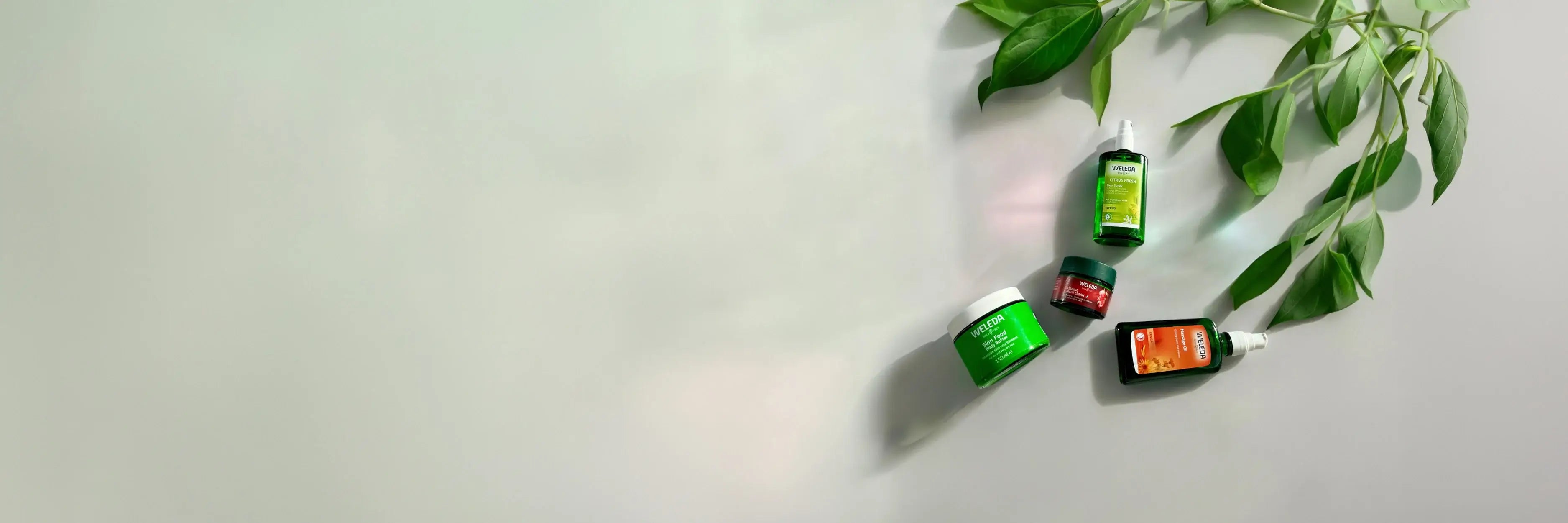 Four Weleda products arranged on a light gray surface with green leaves in the top right corner.