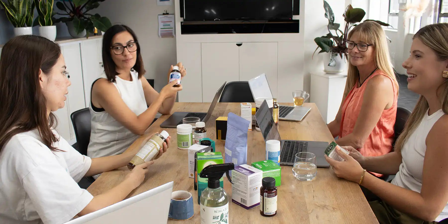 Four women in a meeting room with various items on the table, including bottles and a laptop.
