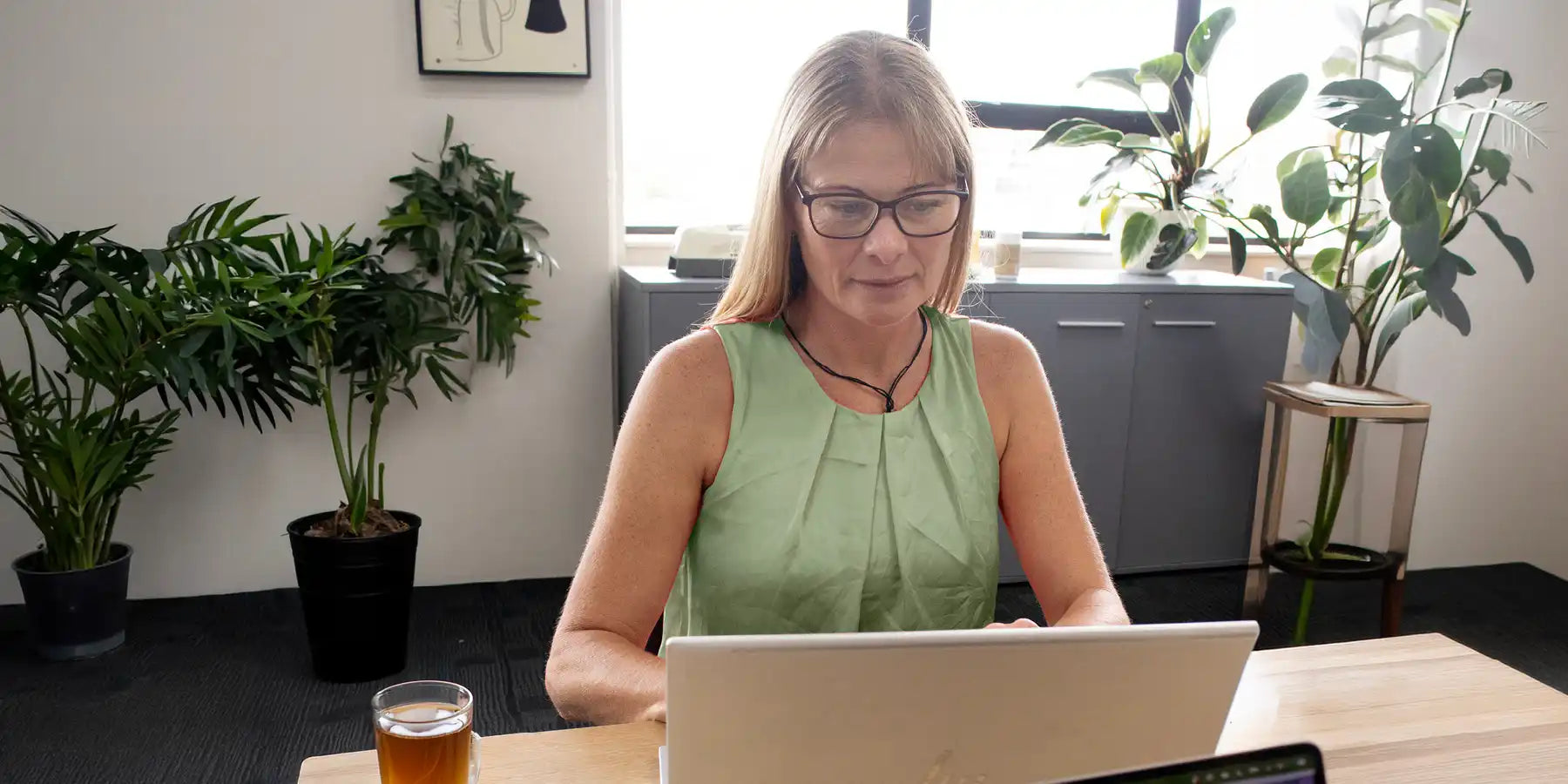 Woman in a green sleeveless top sitting at a desk with plants in the background.