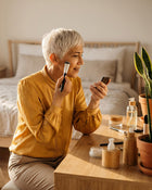 Woman with short white hair applying makeup in a bedroom setting.