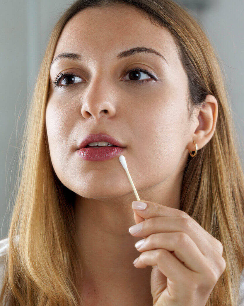 Woman using a cotton swab near her mouth with a neutral background.