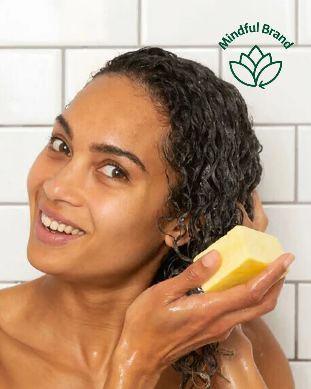A woman with wet curly hair holds a yellow soap bar in a shower setting with white tiled walls. A green 'Mindful Brand' logo is visible in the top right corner.