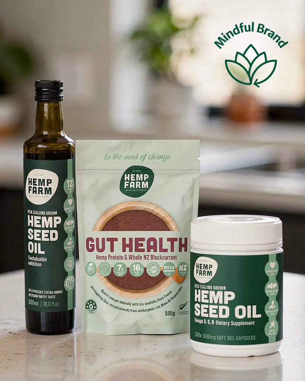 Three Hemp Farm products arranged on a countertop: a dark glass bottle of hemp seed oil, a mint pouch labeled gut health hemp protein powder, and a white tub of hemp seed oil gels. A green 'Mindful Brand' logo appears in the upper right.