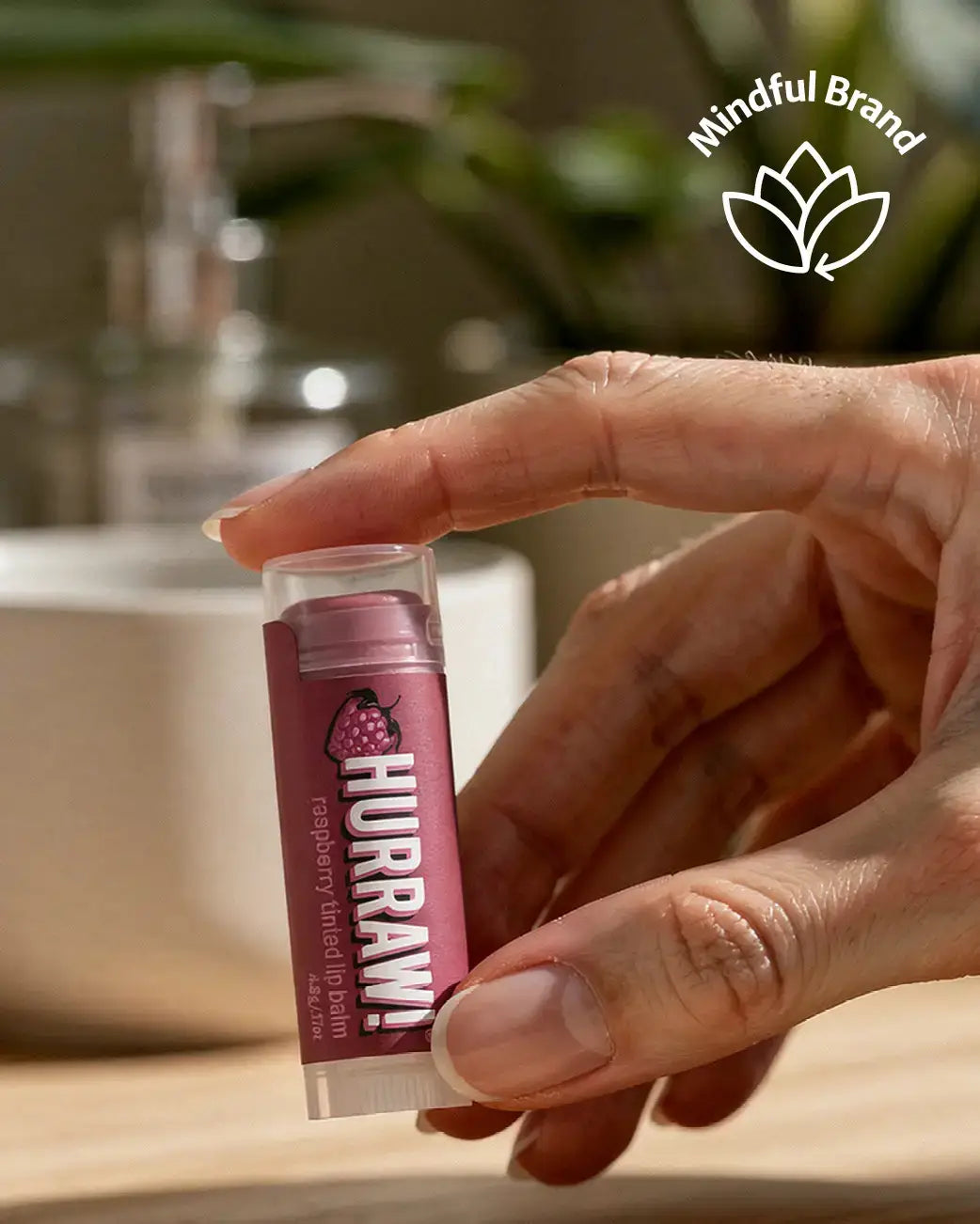 A hand holding a tube of Hurawi Raspberry Hand Lip Balm against a blurred bathroom background with a sink and faucet. A white 'Mindful Brand' logo is visible in the top right corner.