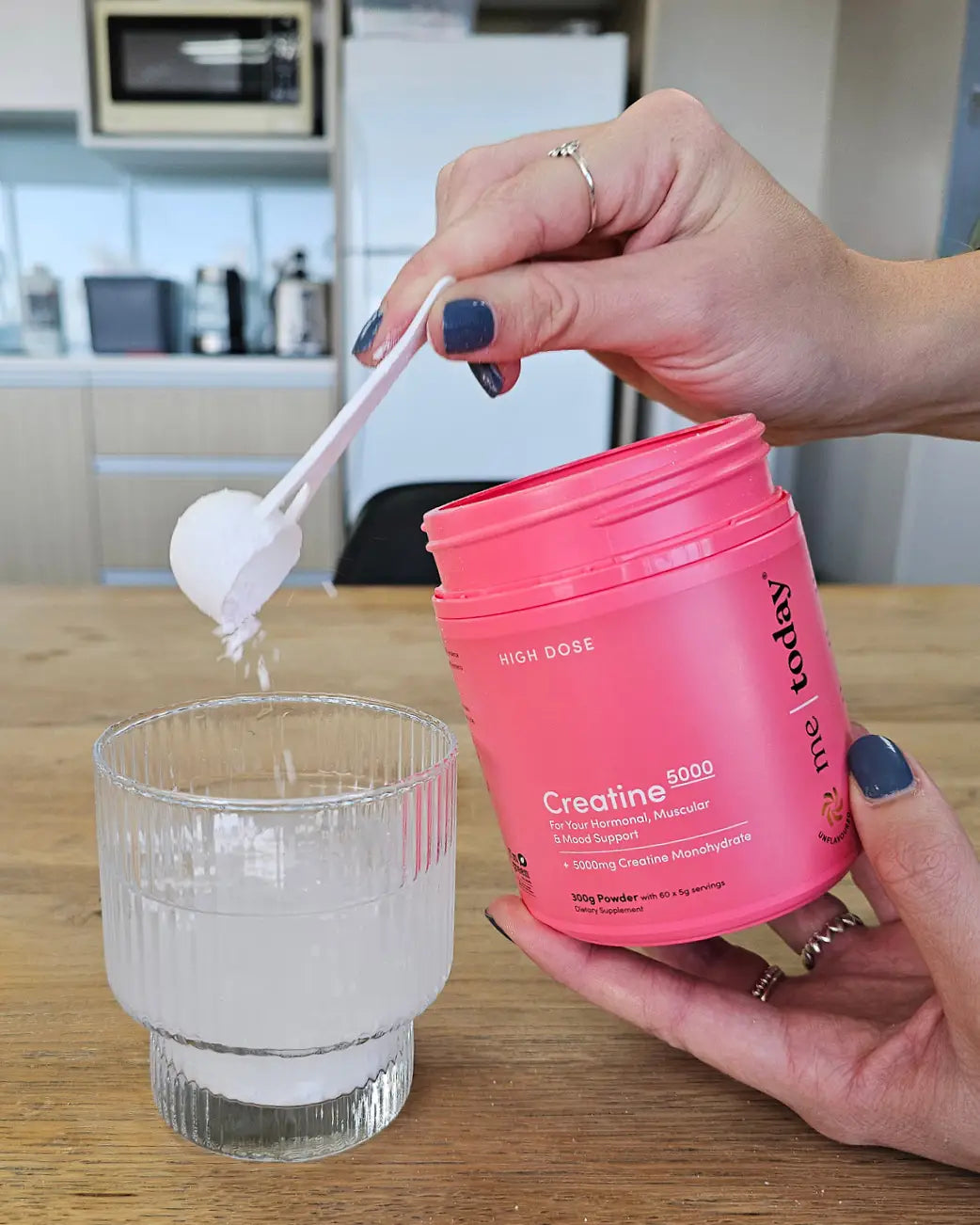 A person's hands pour white creatine powder from a scoop into a clear glass of water, holding a pink tub of High Dose Creatine powder.
