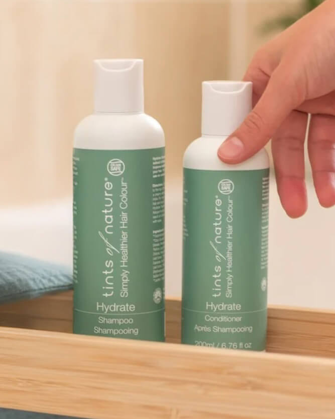 Two bottles of 'Tints of Nature' shampoo and conditioner on a wooden surface with a hand reaching for one bottle.