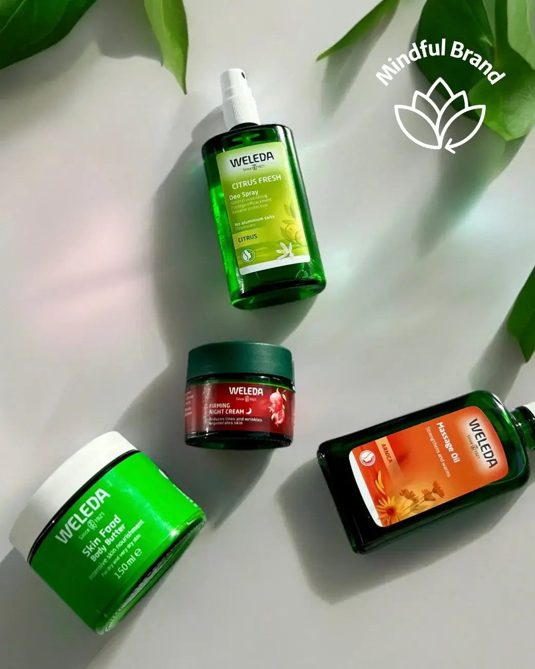 Four Weleda skincare products arranged on a light gray surface with green leaves: a green spray bottle, a red-and-green cream jar, a green cream jar, and a dark green liquid bottle. A white 'Mindful Brand' logo appears in the top right.