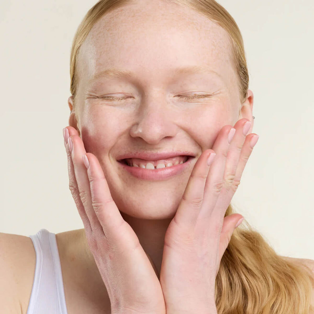 Woman with blonde hair and a white tank top touching her face against a beige background.