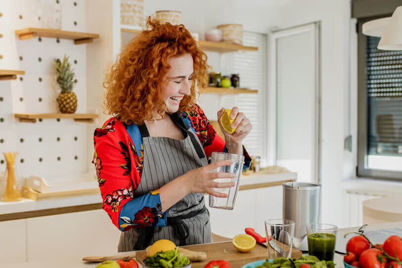 Woman happily squeezing some lemon juice into her blender with fruits and vegetables on a bench.