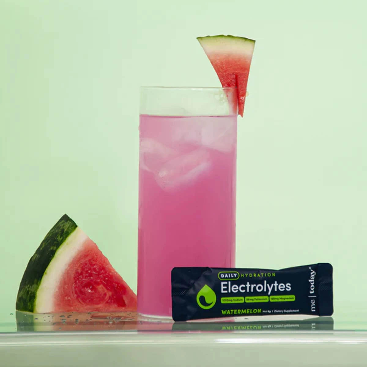 Glass of pink watermelon electrolytes with a sachet sitting next to it and slices of watermelon.